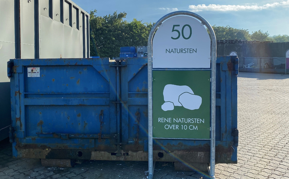 Skilt med teksten Natursten står foran en blå container på en genbrugsplads