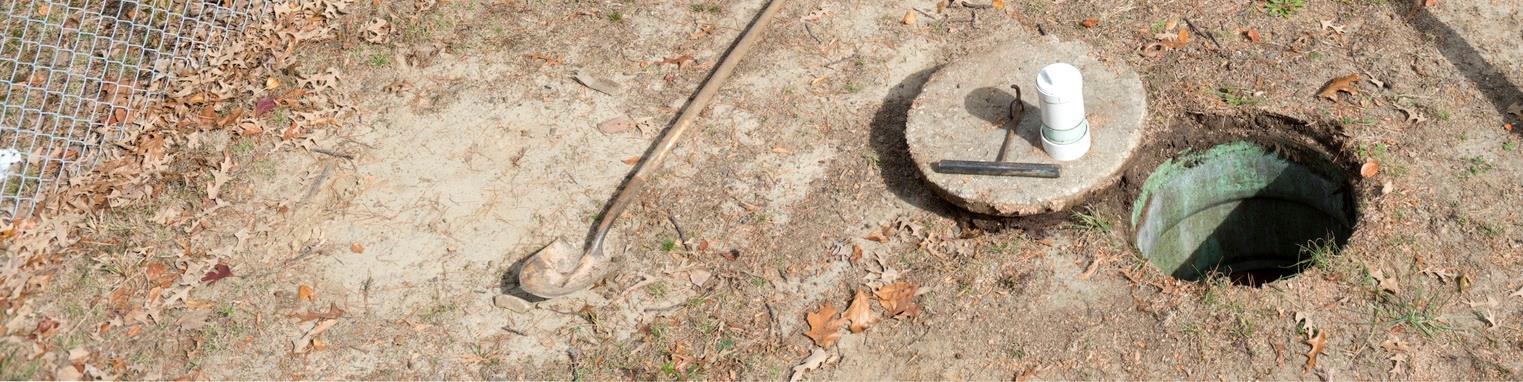 På billedet ser man et hul i jorden og et dæksel, der ligger ved siden af hullet. I venstre side af billedet kan man se noget af et trådhegn. På jorden ligger der en skovl.