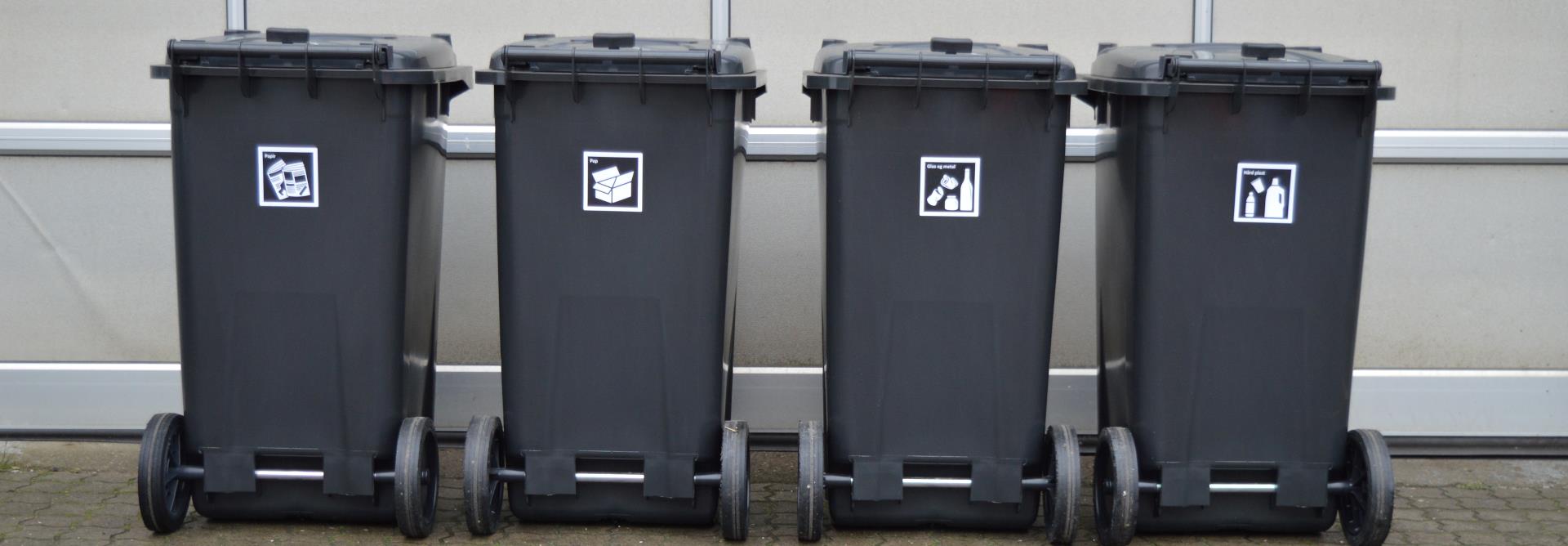 Fire sorte containere står ved siden af hinanden op ad en port. På hver container er der et piktogram, der illustrerer hvad der må komme i. Fra venstre er det papir, derefter pap, så glas og metal og den sidste container er til hård plast.