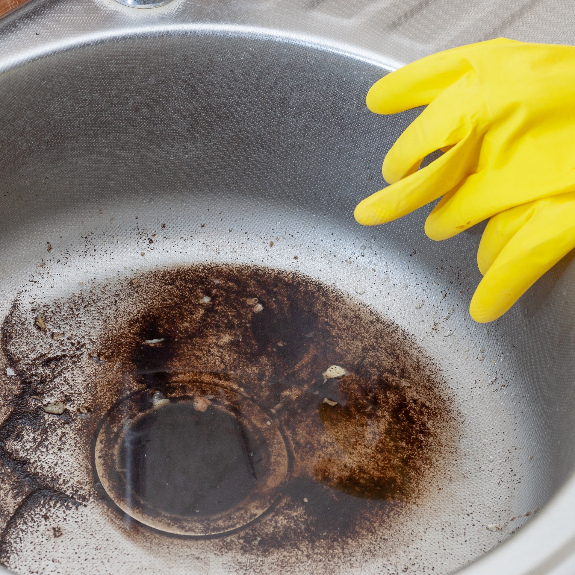 På billedet ser man en rund metalhåndvask. I bunden af vasken ligger der noget flydende fedt og snavs. På kanten af vasken ligger der nogle gule gummihandsker.