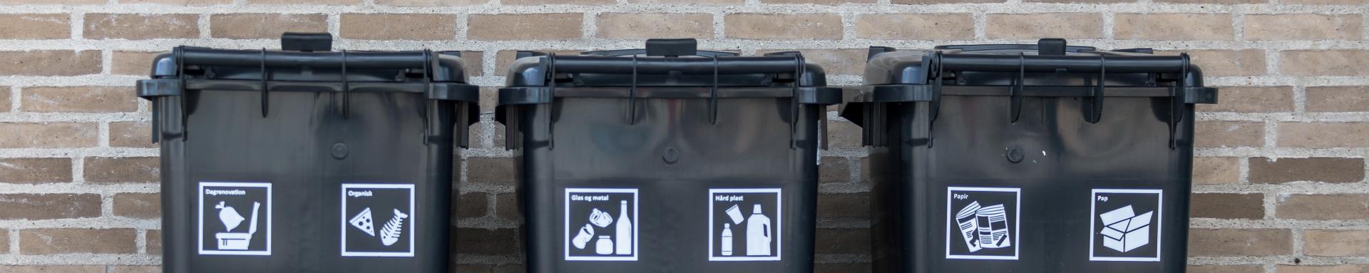 Tre sorte affaldscontainere står op ad en lysebrun murstensvæg. På hver containere er der to hvide piktogrammer, som hver især illustrerer hvilken type affald, der må komme i containeren.  