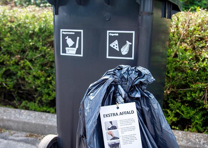En sort affaldscontainer med en sort affaldssæk stående foran. På affaldssækken er der bundet et mærkat fast. På mærkatet står der ekstra affald.