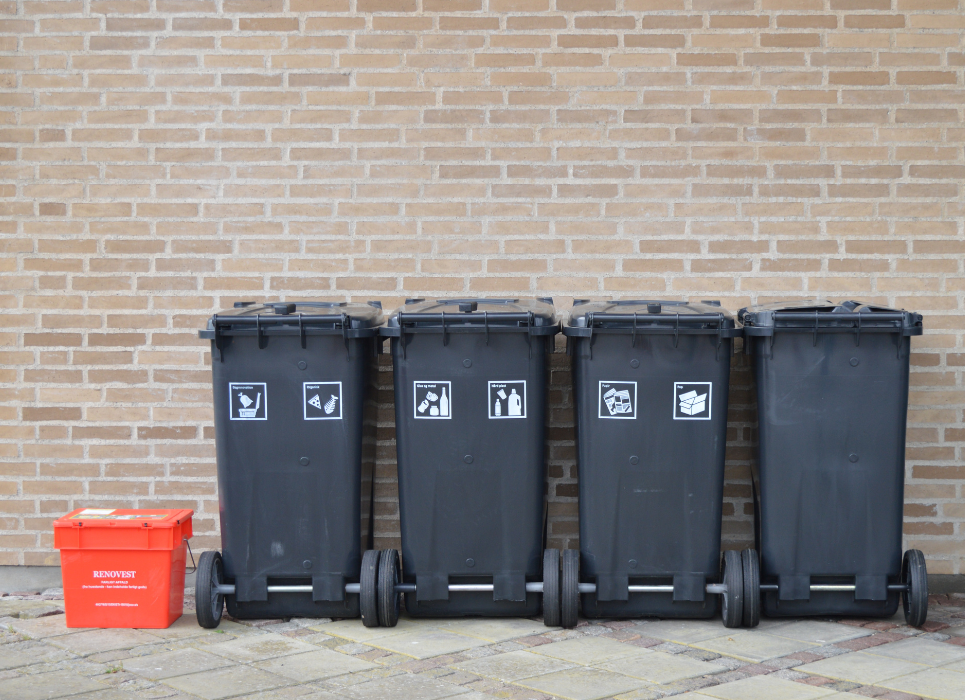 Fire sorte affaldsbeholdere og en rød miljøkasse står på stribe op ad en gulbrun murstensvæg