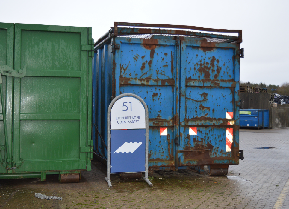 En grøn og en blå container på en genbrugsplads med et skilt foran hvor der står eternitplader uden asbest