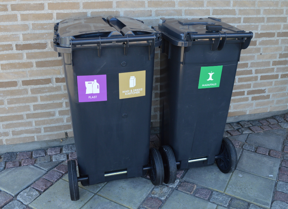 To sorte affaldsbeholdere står op ad en murstensvæg
