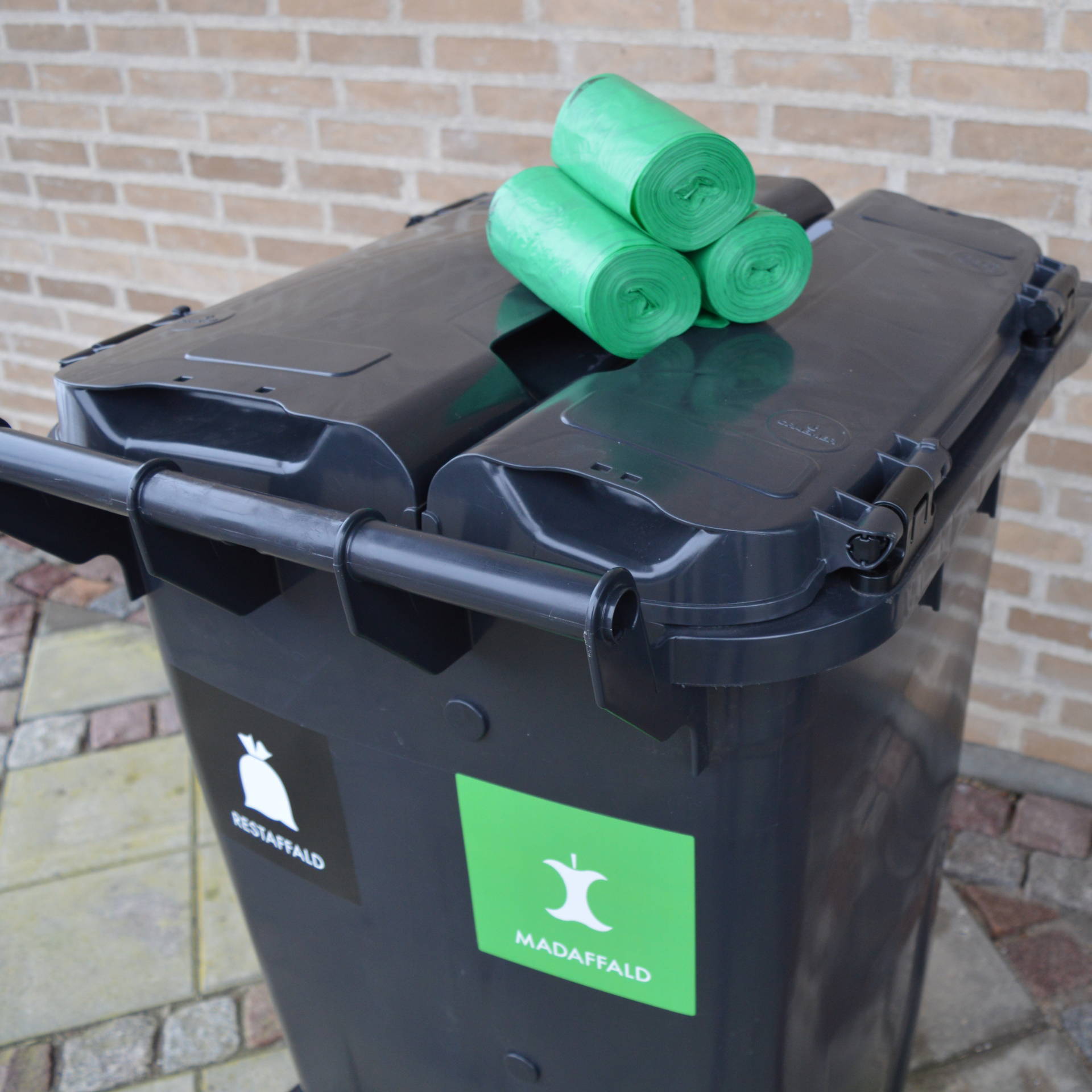 tre ruller grønne affaldsposer ligger ovenpå låget af en sort affaldsbeholder